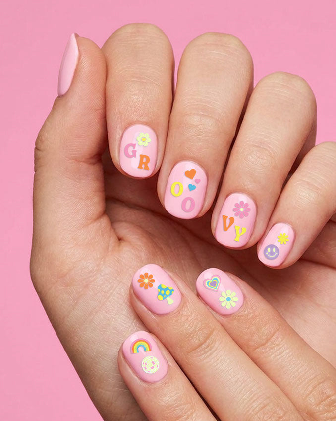 Hand with pink nails wearing groovy nail stickers including GROOVY letters, daisies, a heart, and a rainbow