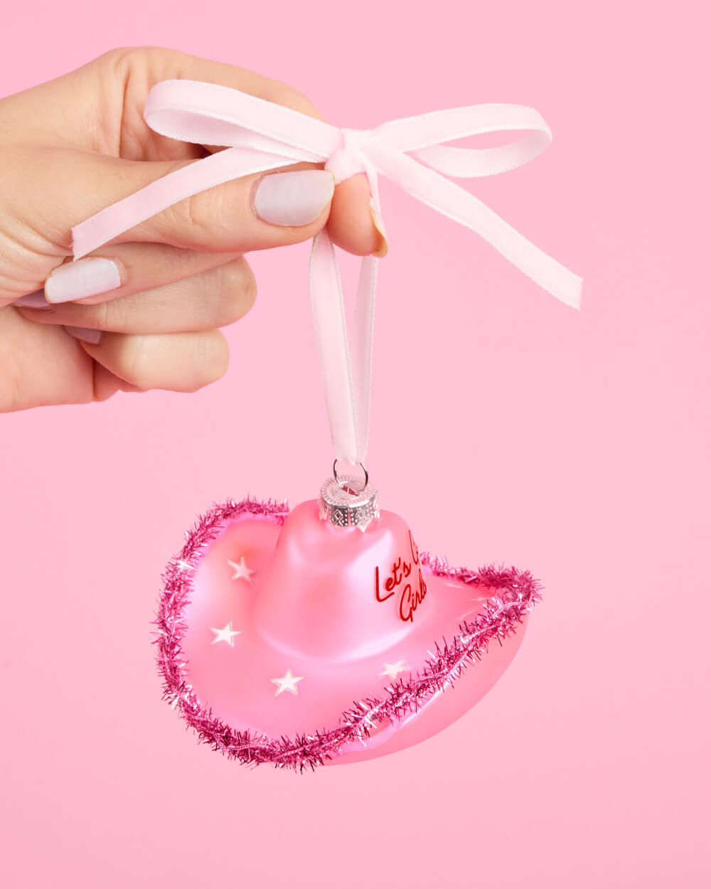 Hand holding a pink cowgirl hat Christmas ornament by its pink ribbon loop on a pink background