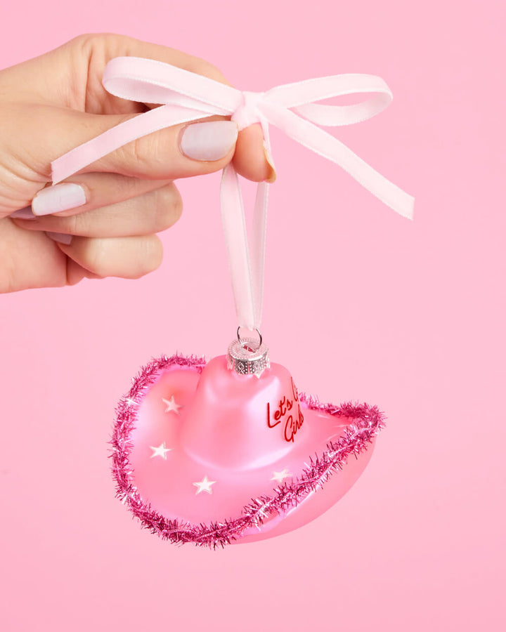 Hand holding a pink cowgirl hat Christmas ornament by its pink ribbon loop on a pink background