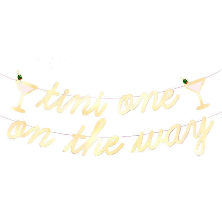 Pre-strung gold foil tini one on the way banner with a martini glass icon at each end on a pink wall