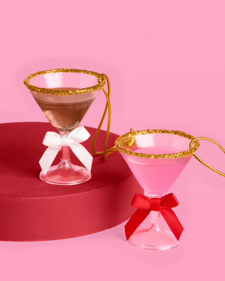 Two mini martini ornaments, espresso with a white bow and pink with a red bow, gold glitter rims, on a red riser