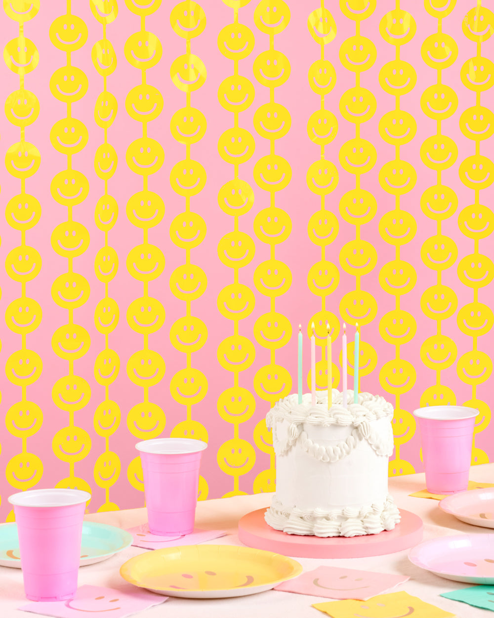 Yellow smiley face curtain behind a white birthday cake with pink cups and yellow plates