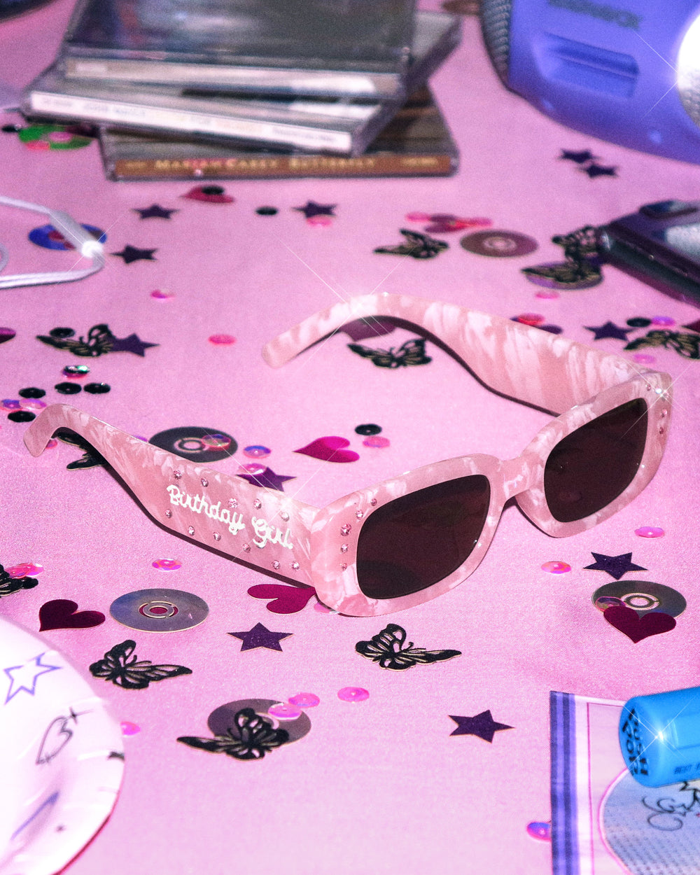 Pink marble Birthday Girl sunglasses with brown lenses on a pink table scattered with CDs and confetti