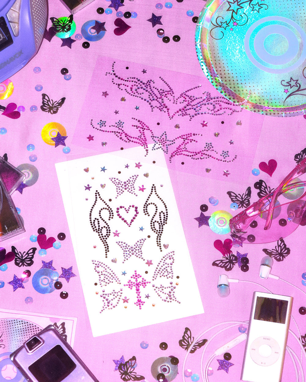 Sheet of black and pink Y2K body gems with butterflies, hearts, and a cross on a table of CDs and confetti