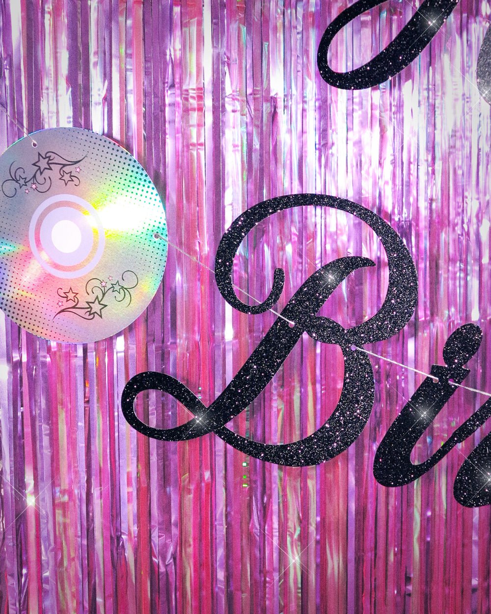 Close-up of black glitter Birthday lettering and an iridescent foil CD on the Y2K banner against purple fringe