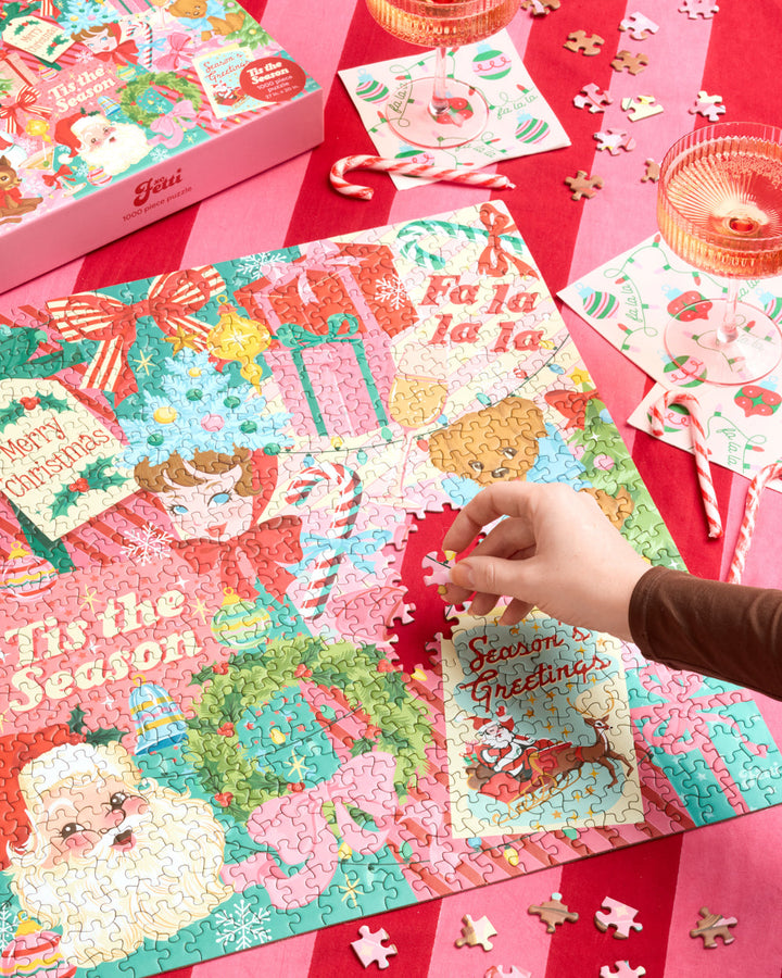 Hand placing a piece into the Pinkmas Christmas puzzle on a red-and-pink striped table.