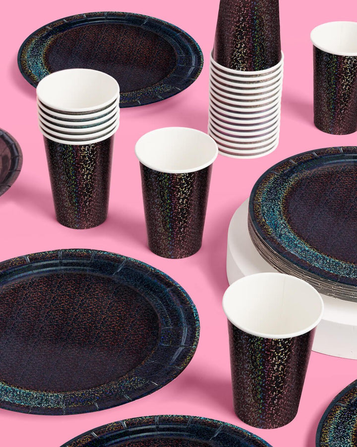Black iridescent paper cups laid out on a pink surface beside dark speckled party plates