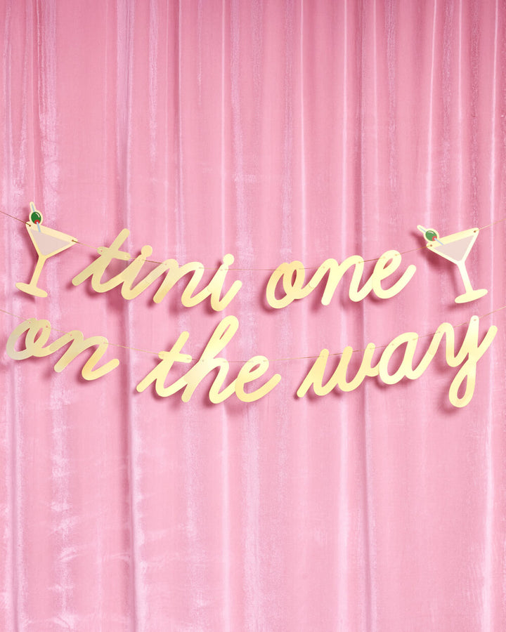 Tini one on the way banner in gold foil script hung over a pink velvet curtain for a cocktail baby shower