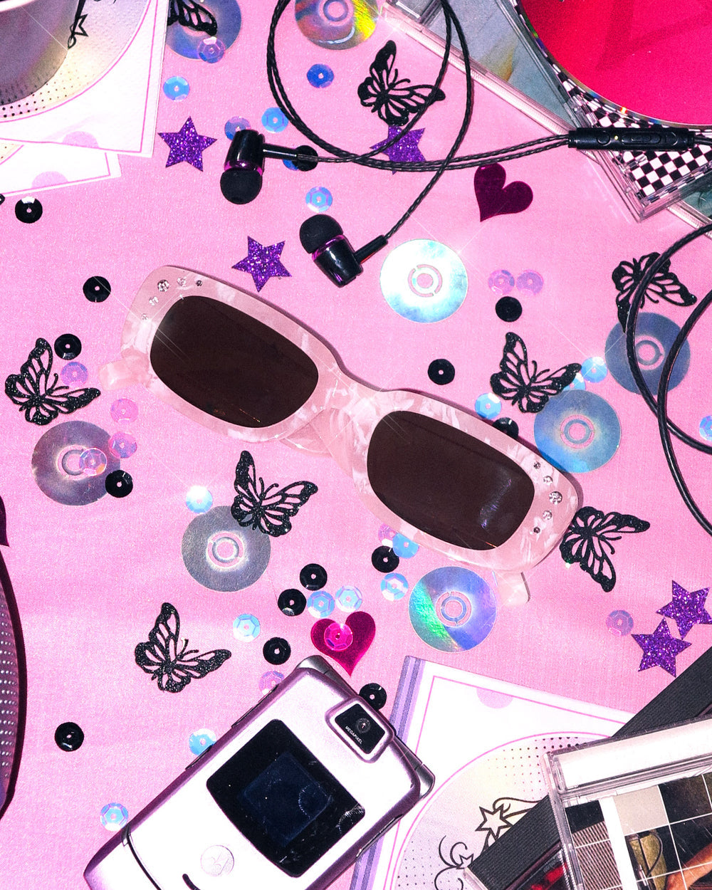 Pink rhinestone birthday sunglasses with a flip phone, butterflies, and CDs on a pink table