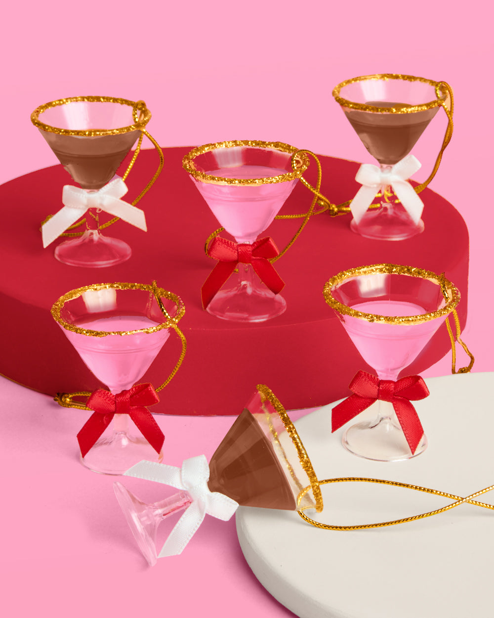 Six mini martini Christmas ornaments, three pink and three espresso, with gold glitter rims on red and cream risers