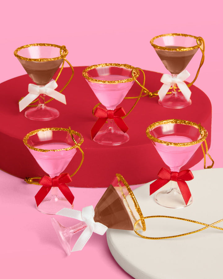 Six mini martini Christmas ornaments, three pink and three espresso, with gold glitter rims on red and cream risers