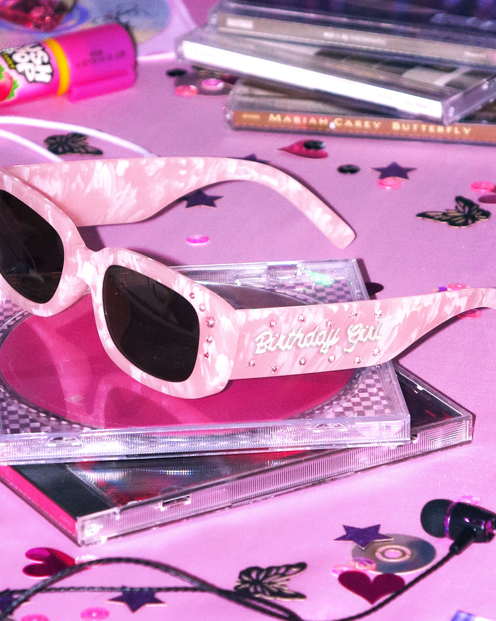 Pink marble birthday sunglasses with rhinestones resting on a CD case with star confetti