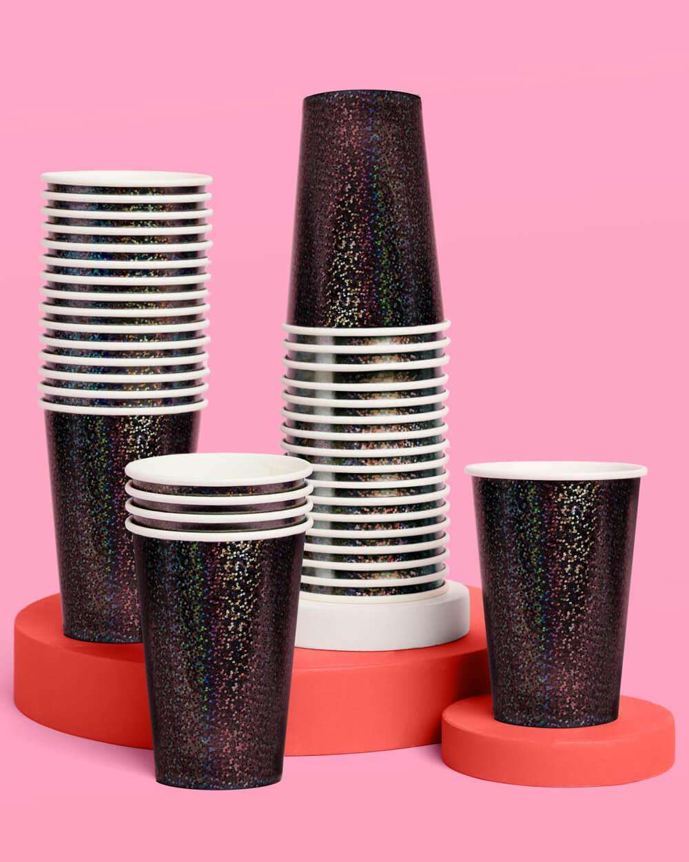 Black iridescent paper cups stacked on orange risers against a pink background