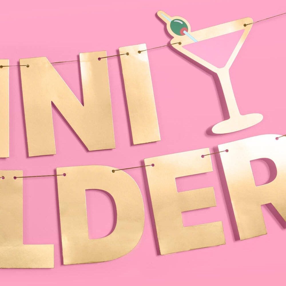 Detail of a pink martini glass with green olive pick beside gold foil lettering on the birthday banner string