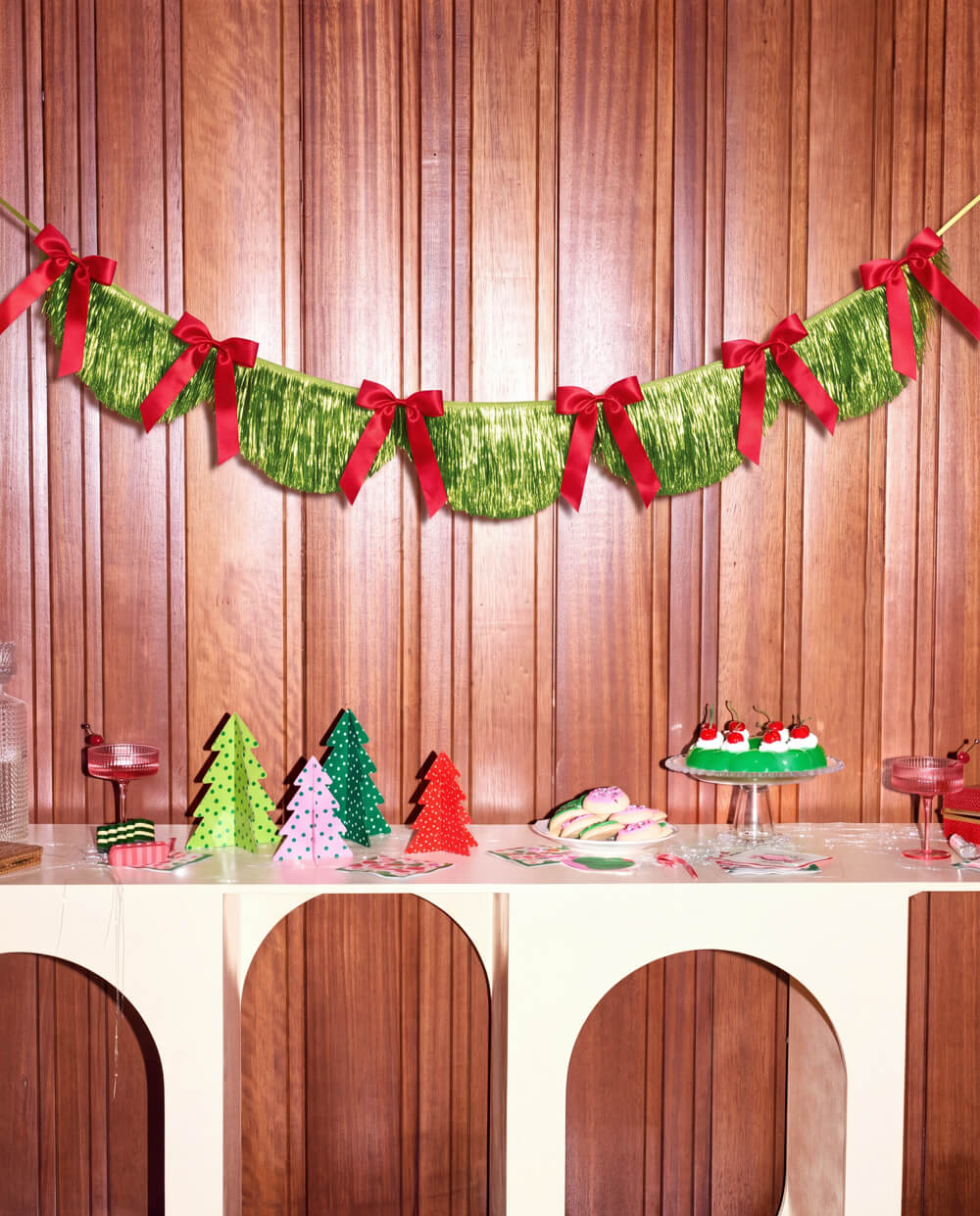 Green fringe banner with red bows hung across a wood wall above a dessert table