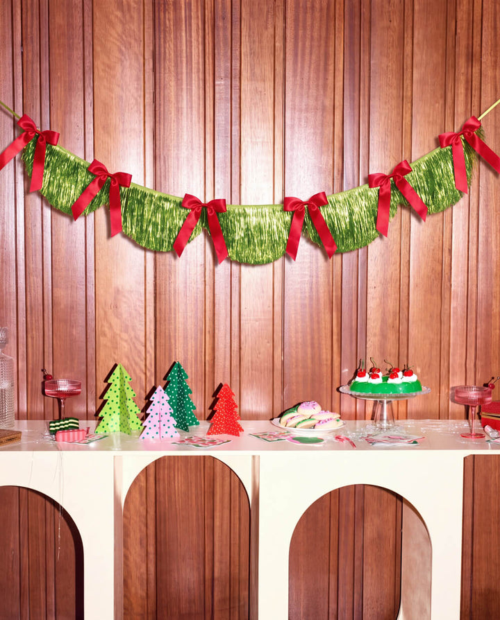 Green fringe banner with red bows hung across a wood wall above a dessert table