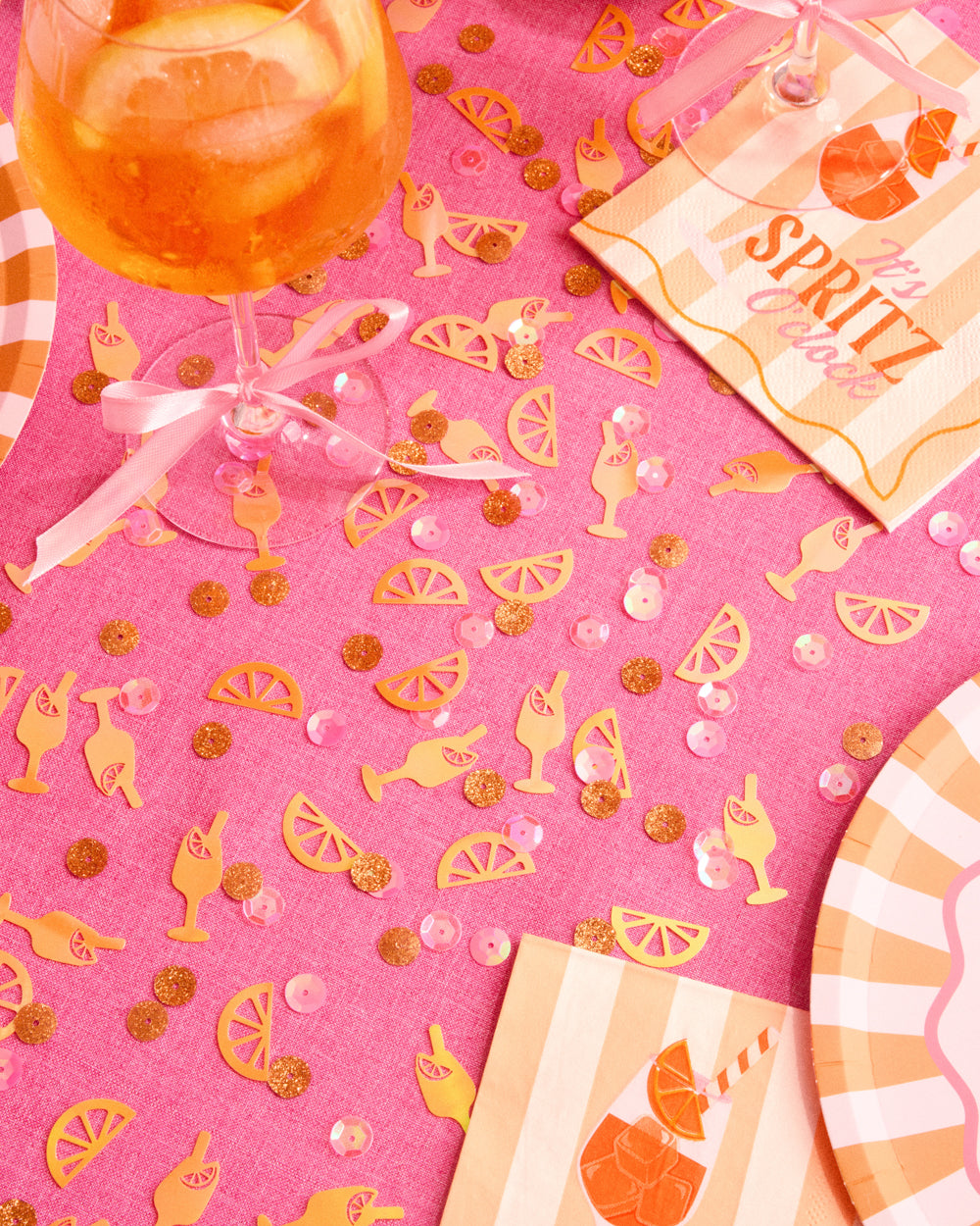 Spritz party confetti scattered on a pink table with spritz cocktails and orange striped plates