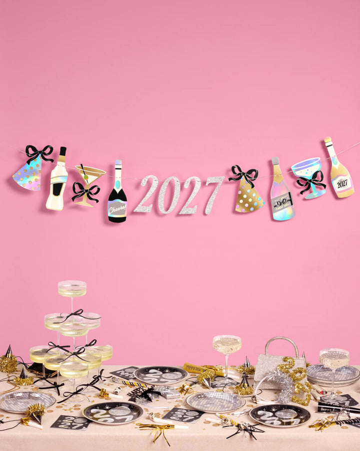 2027 New Year banner with foil bottles, cocktail glasses, and party hats above a festive table.
