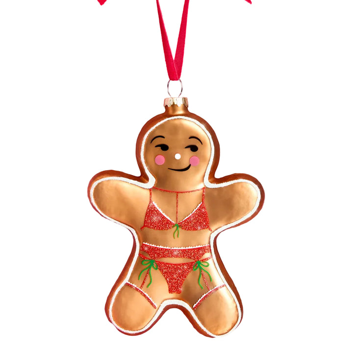 Gingerbread girl glass Christmas ornament with a winking face, pink cheeks, and a red glitter bikini with green bows, hanging from a red velvet ribbon with gold hardware.