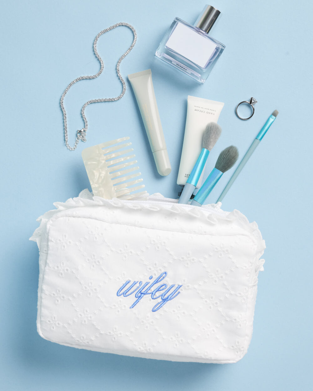 White eyelet cosmetic bag with blue wifey embroidery, styled with makeup brushes, perfume, and jewelry.