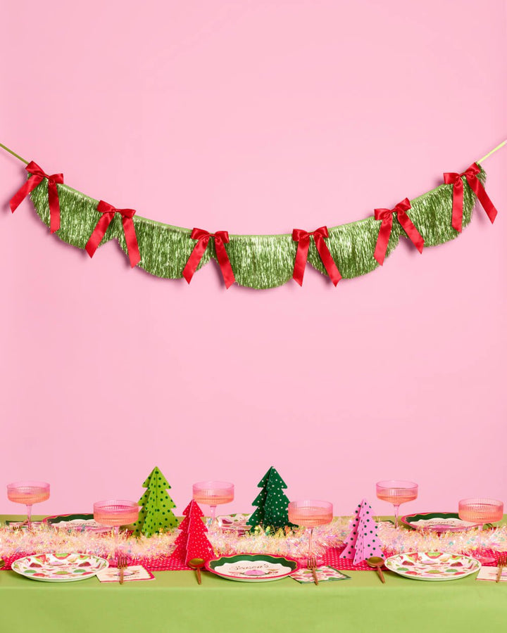 Green fringe banner with red bows hung on a pink wall above a Christmas table
