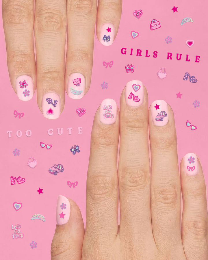 Two hands wearing pink nail stickers with bows, hearts, and handbags on a pink sheet reading TOO CUTE and GIRLS RULE