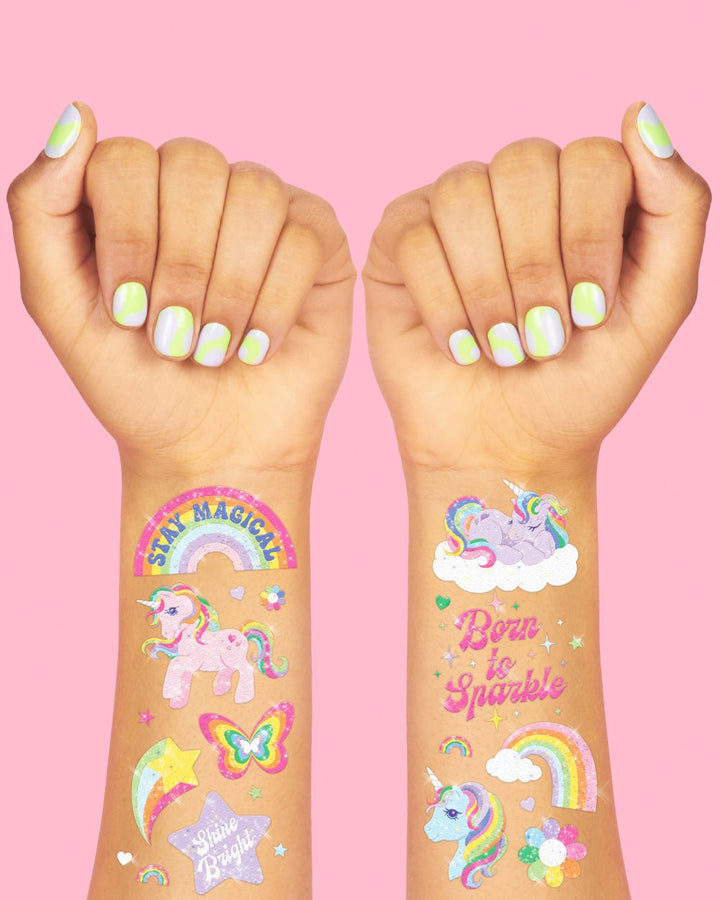 Two arms wearing unicorn temporary tattoos: Stay Magical rainbow, unicorn on a cloud, butterfly, and Born to Sparkle