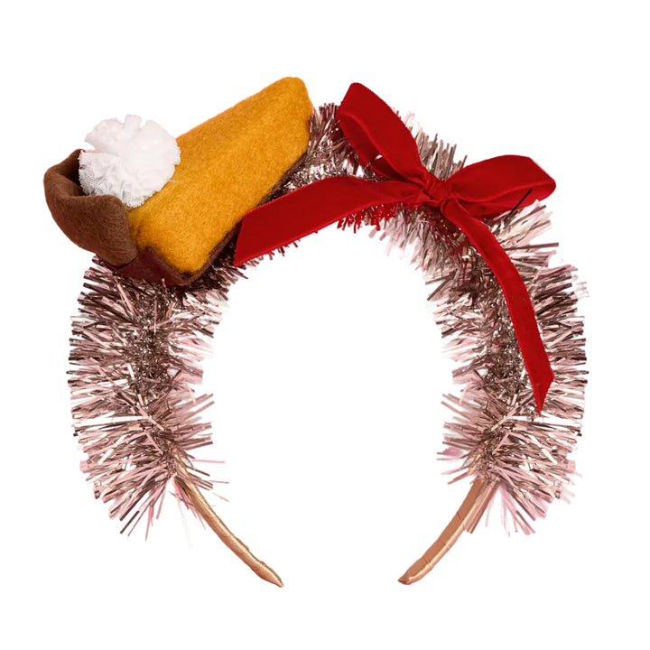 A novelty headband styled like a slice of pie, featuring a metallic silver tinsel arch with a large red ribbon bow, a mustard-yellow felt slice, a brown crust edge, and a white whipped cream pom-pom on a pink background.