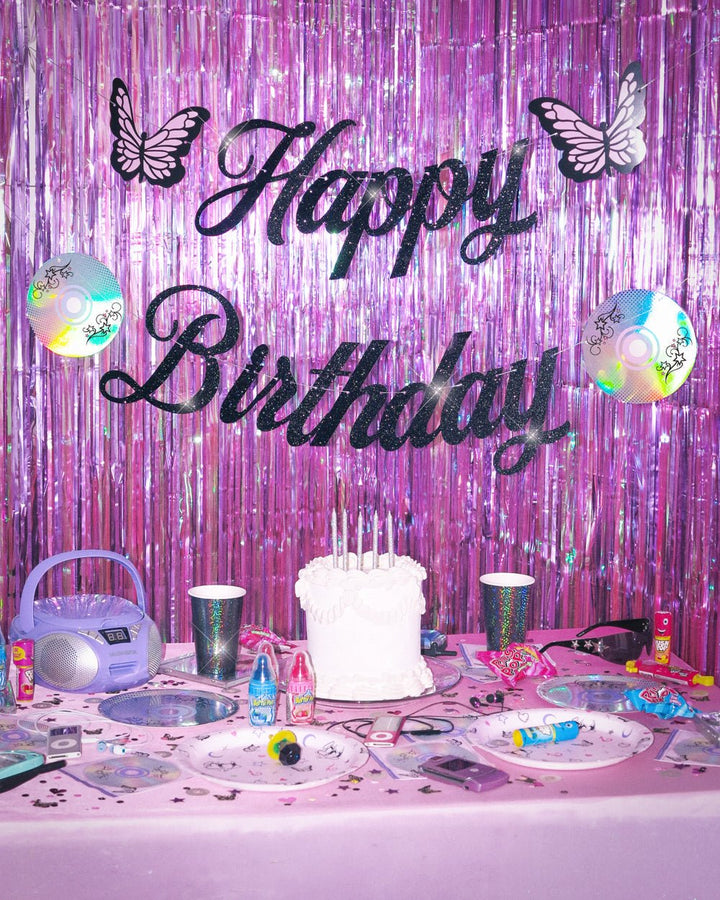 Y2K Happy Birthday banner in black glitter script with butterflies and iridescent CDs over a purple fringe curtain and cake table