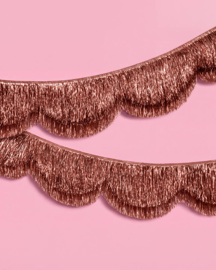 Two strands of rose gold scalloped fringe garland draped across a pink background