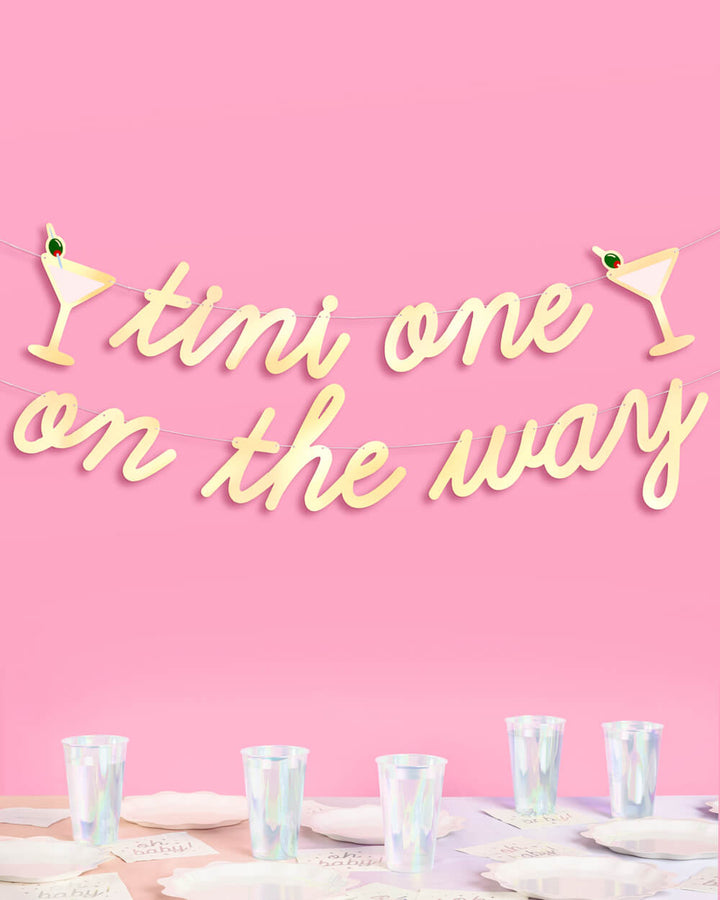 Gold foil tini one on the way baby shower banner with martini glass icons hung above a table of iridescent cups