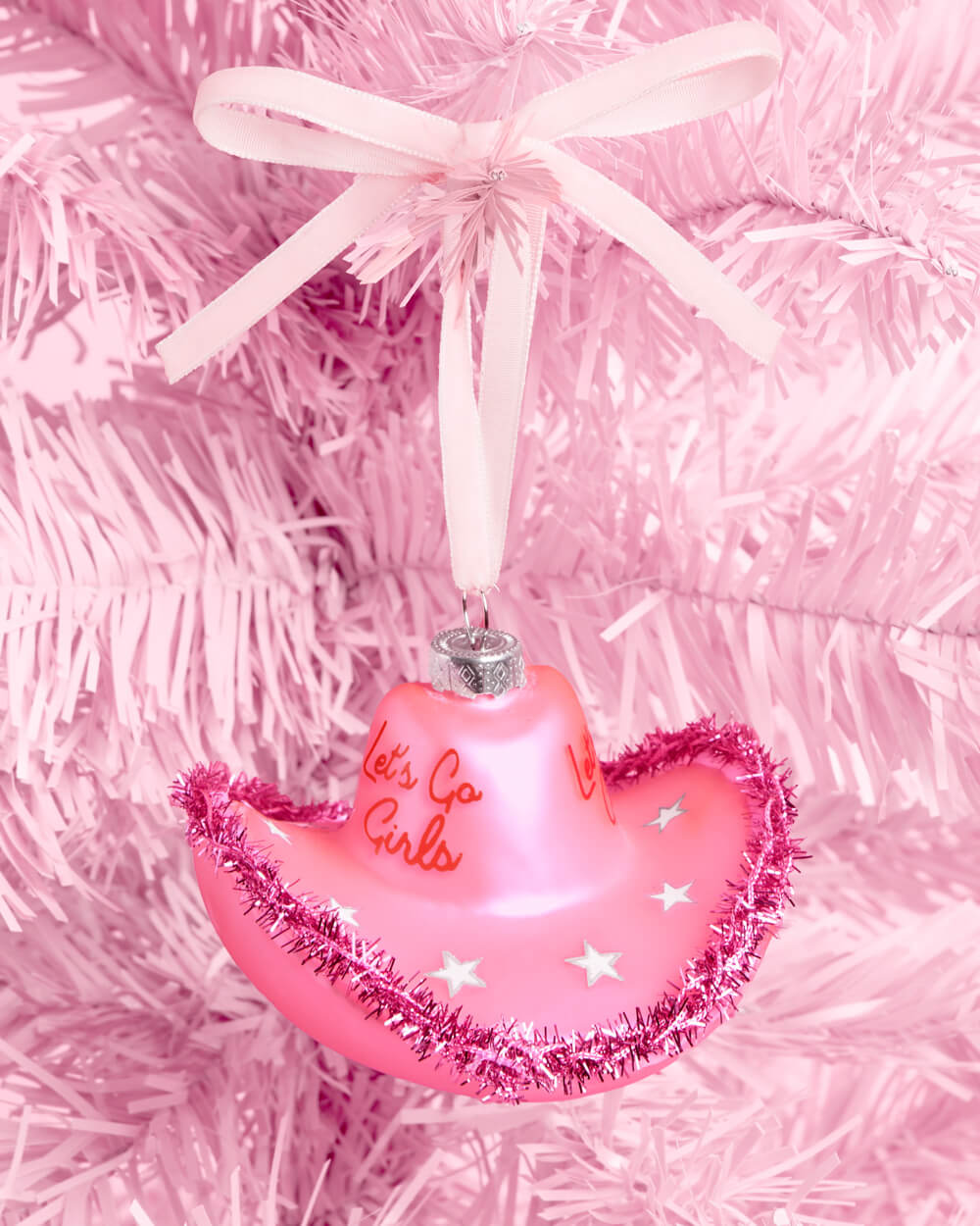 Pink glass cowgirl hat ornament with pink tinsel trim and a ribbon bow hanging on a pink Christmas tree