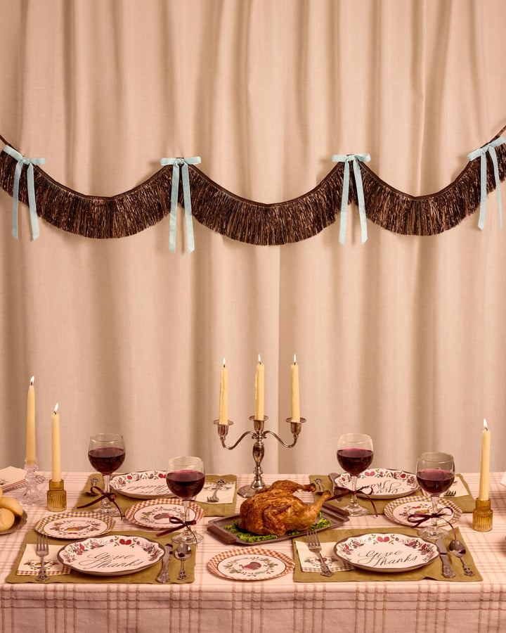 Brown fringe backdrop with light blue velvet bows behind a Thanksgiving table set with a turkey, candles, and wine glasses