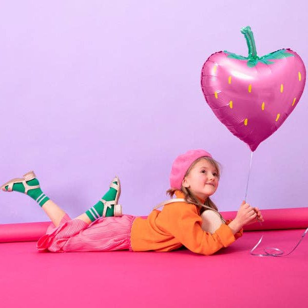 Child in a pink beret holding the pink strawberry foil balloon against a lilac and pink backdrop