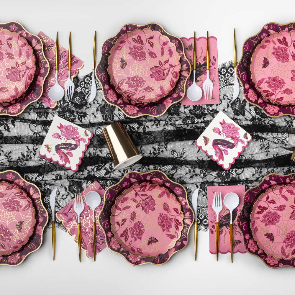 A styled flat-lay of a pink and magenta floral tableware set with gold-toned cutlery, white napkins, and a black-and-white lace runner on a light gray background.