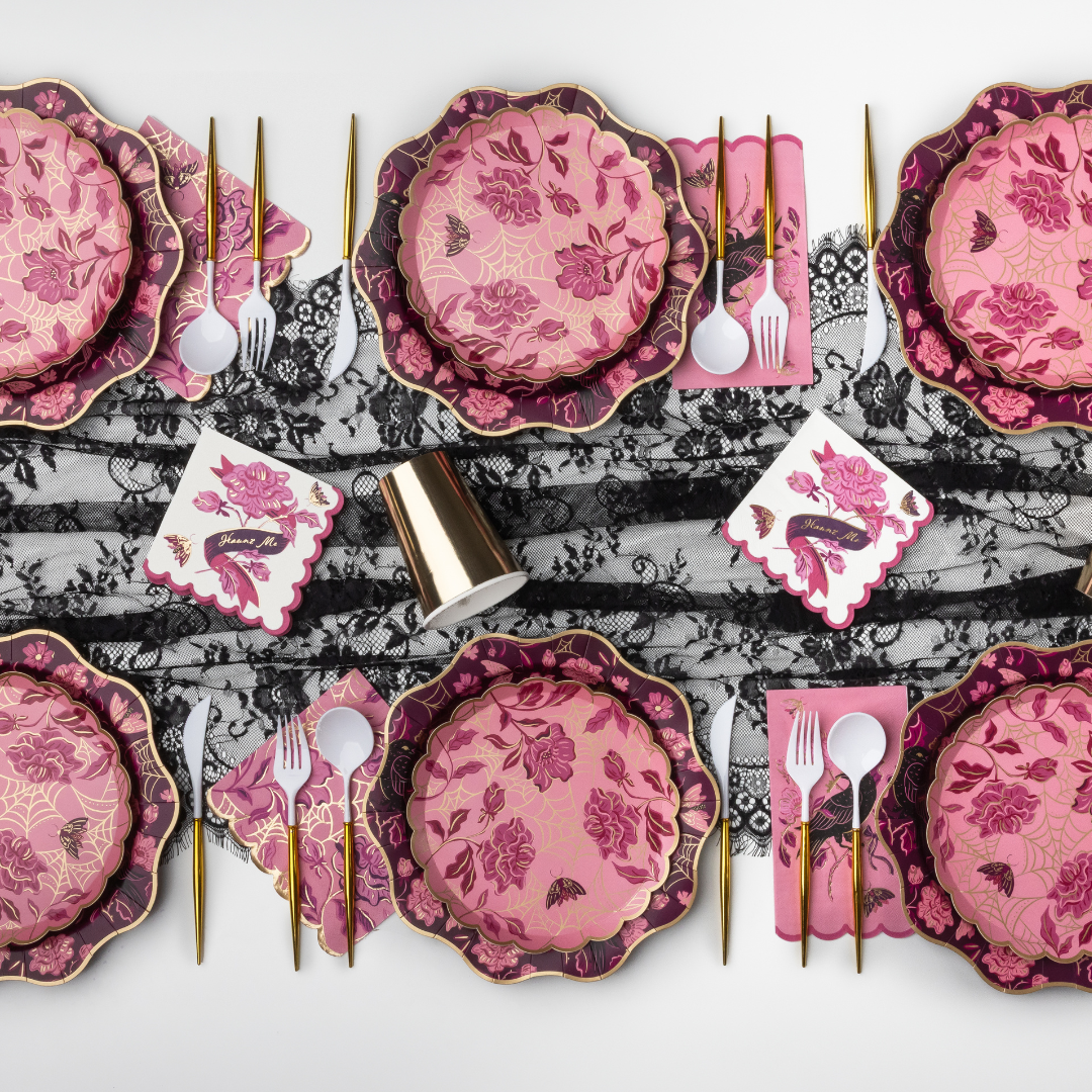 A styled flat-lay of a pink and magenta floral tableware set with gold-toned cutlery, white napkins, and a black-and-white lace runner on a light gray background.