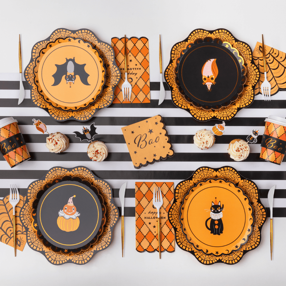 A Halloween-themed table setting featuring four scalloped plates with bat, witch, pumpkin, and cat illustrations, arranged with orange patterned napkins, gold forks, striped table mats, and small cupcakes on a white background.