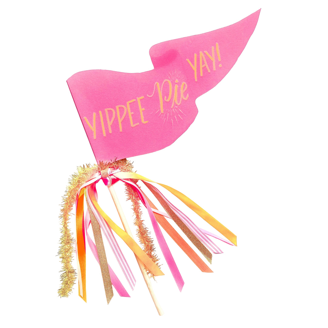 A pink paper tail party accessory with gold foil lettering that reads 'YIPPEE Pie YAY!' and dangling ribbon streamers in pink, orange, gold, and white on wooden sticks, photographed on a white background.